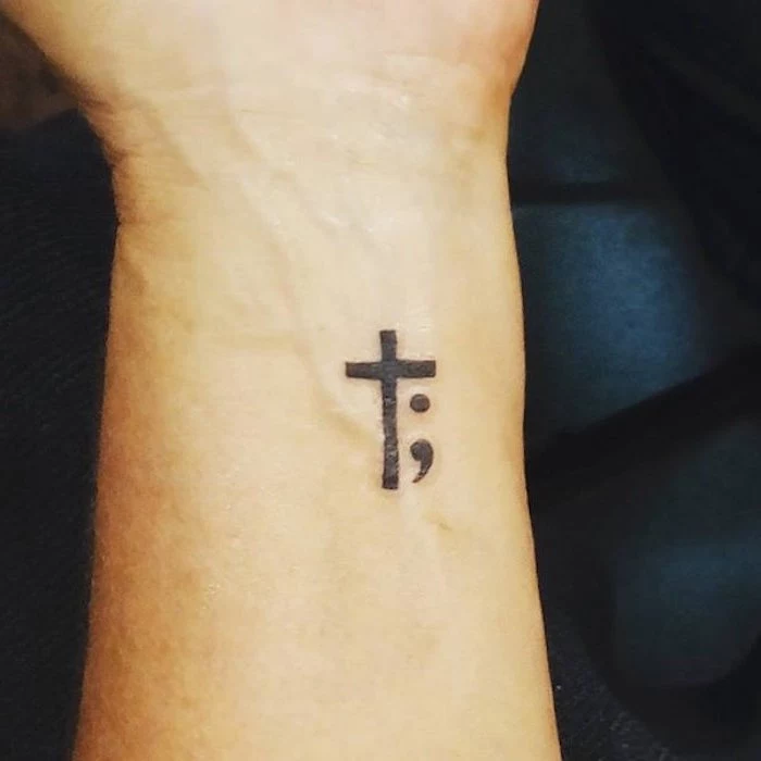 cross tattoo in black, accompanied by a semicolon, near a person's wrist, minimalistic and simple design, tattoo with religious symbolism