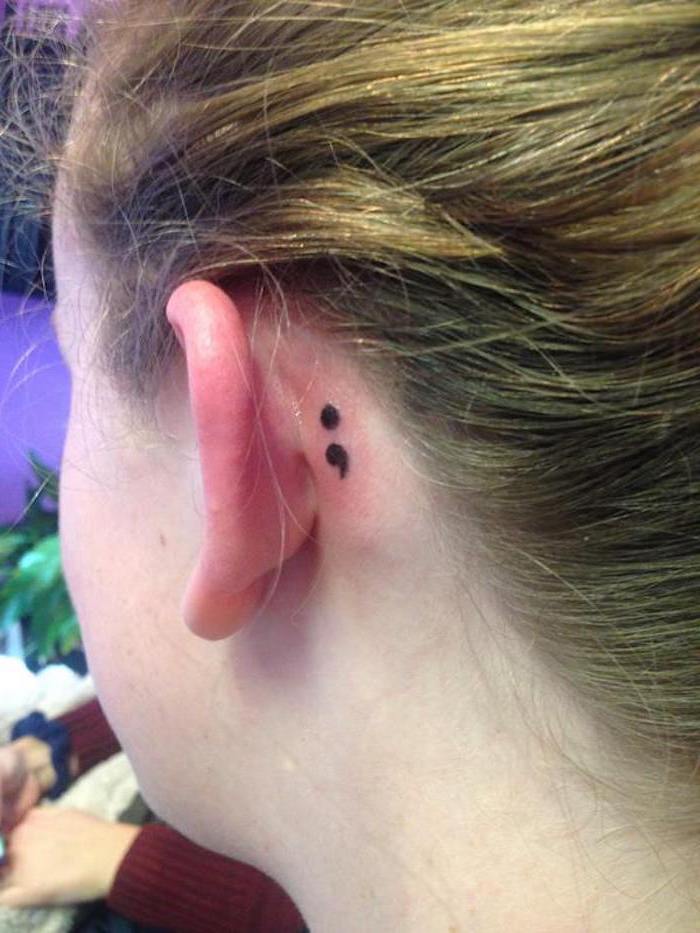 behind the ear tattoo, of a small black semicolon, what does a semicolon tattoo mean, on a pale girl, with blond hair tied back