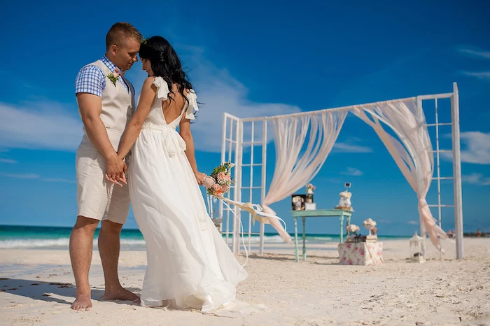 chequered shirt in white and blue, combined with off-white vest and shorts, worn by groom, holding hands with bride in white gown, on beach with fine sand