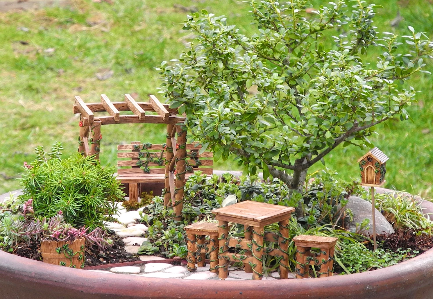 gazebo and bench, table and two stools, bucket and bird house, all made from wood-like material, in a large pot, containing pebbles and plants, and a green bonsai tree