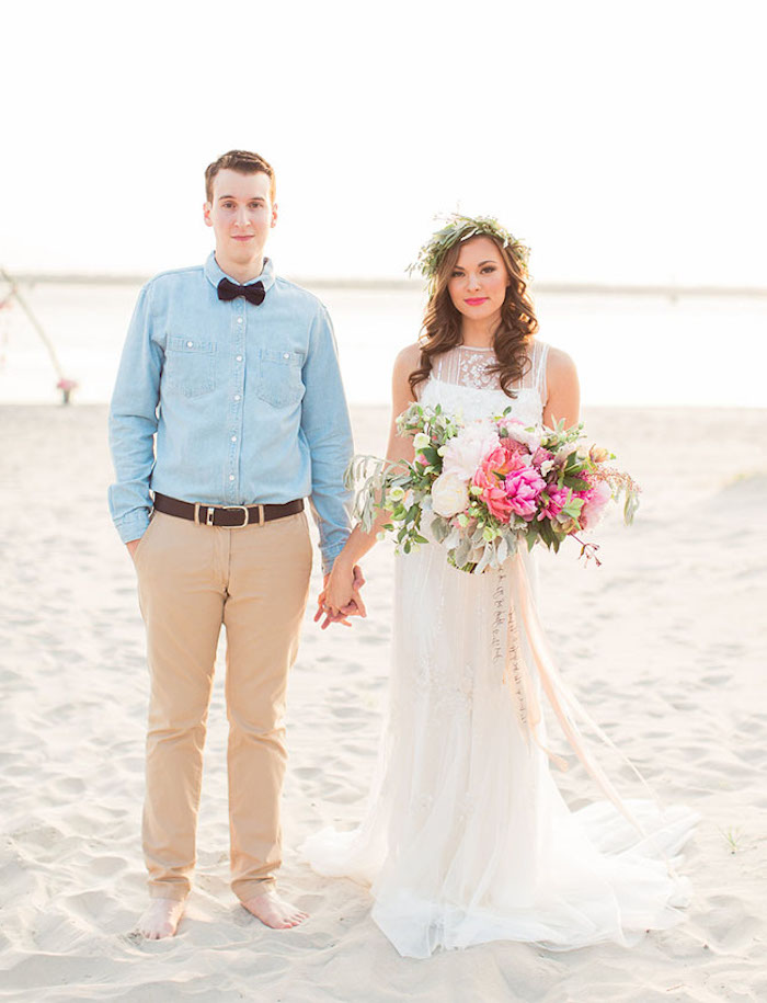 groom in a pale blue shirt, black bowtie and beige pants, holding hands with brunette bride, in a white lace beach wedding dress, holding large multi colored bouquet