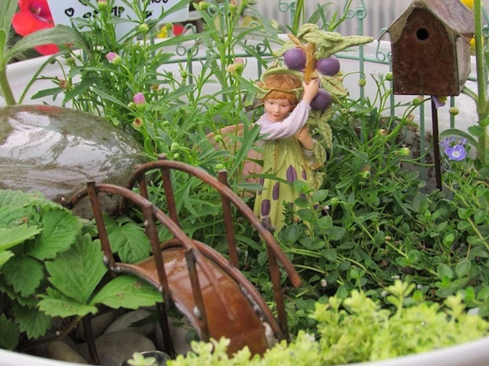 brown metal bridge figurine, placed inside a small garden, near fairy statuette, in white and green dress, and holding a branch with purple berries, fairy garden images, miniature birdhouse and stones