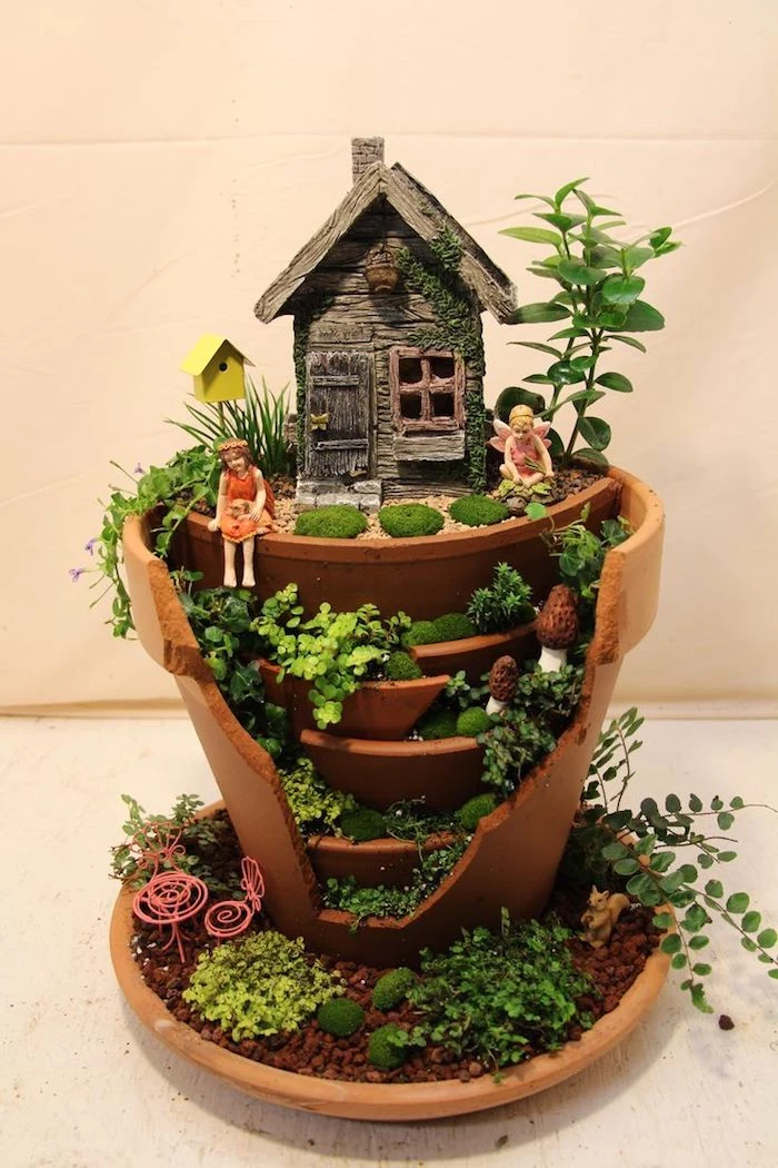 layered-effect broken ceramic pot, decorated with a tiny house, miniature birdhouse and two fairy figurines, fairy garden images, several different small green plants