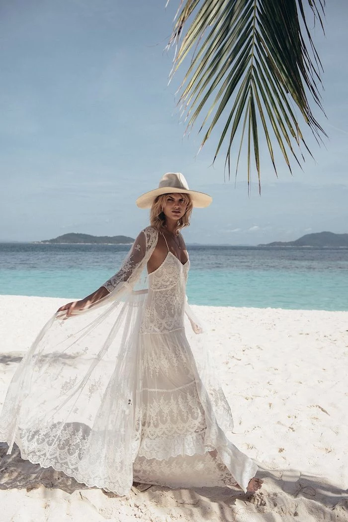 sheer oversized lace cardigan, worn over an embroidered lace maxi dress, casual beach wedding dresses, by blonde model, with yellow sun hat, walking on a sandy white beach
