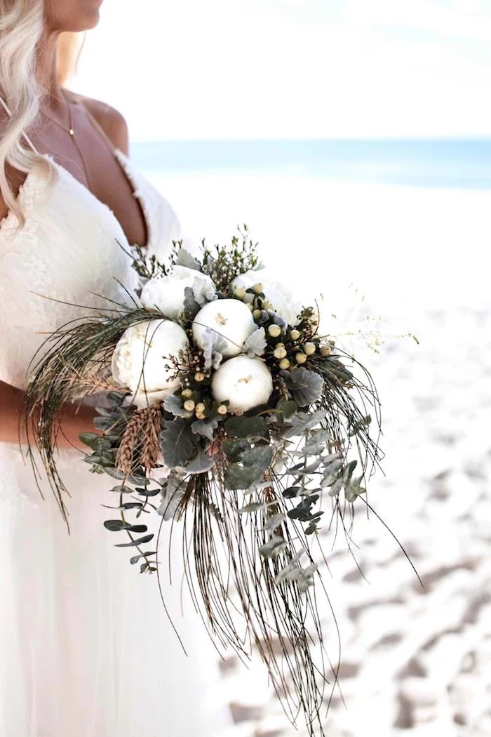 bouquet with white peonies, and various green plants, held by tanned blonde bride, wearing long white gown, and standing on a sandy shore near the sea, beach weddings in florida, white fine sand