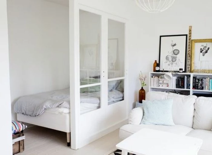 sleeping area with small bed, half-separated from the living room area, through a white partial wall, with glass inserts, apartment design, off-white sofa and bookshelves nearby