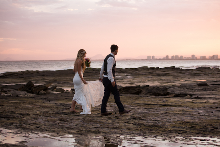 pink sunset sky, beach weddings in florida, barefoot bride in long backless white gown, and groom in black white shirt, black pants and vest, walking on a stony sea shore