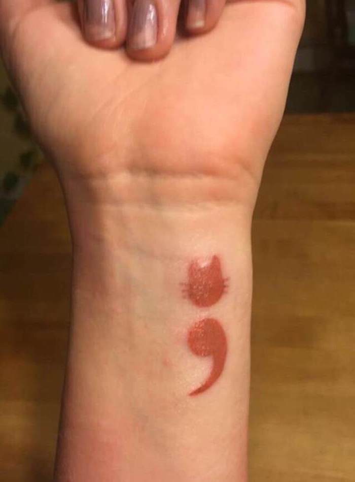 cat shape made from a semicolon, with whiskers and ears, tattooed in red, on the inside of a person's wrist, suicide awareness tattoo 