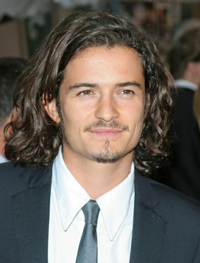 orlando bloom smiling, with medium length, brunette curly hair, combed over to one side, curly hairstyles, short mustache and goatee, formal suit with white shirt, and black shiny tie