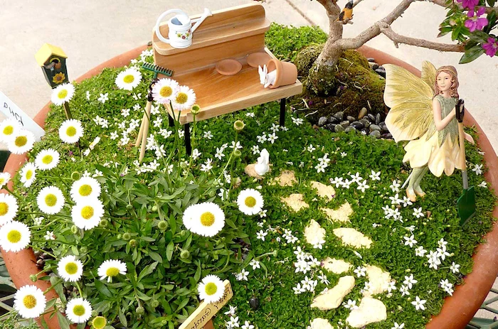 bench-shaped miniature wooden figurine, inside a large ceramic pot, with daisies and tiny white flowers, fairy garden images, decorated with a yellow and green fairy figurine, holding a shovel