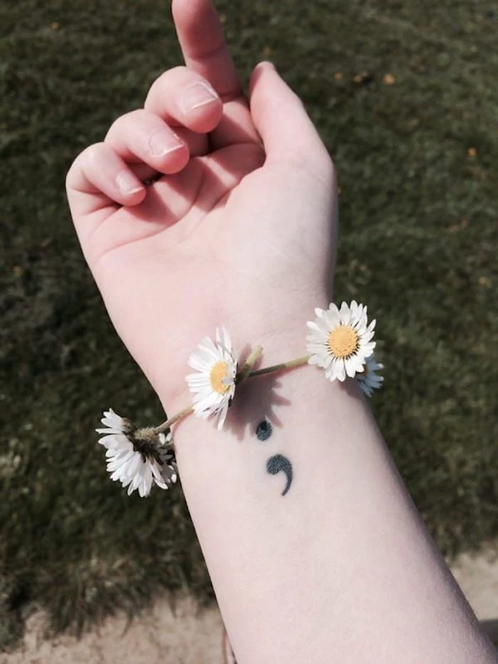 bracelet made from fresh daisies, woven in a chain, worn on the wrist of a pale person, with short nails and a small black tattoo, semicolon tattoo meaning, green field in the background