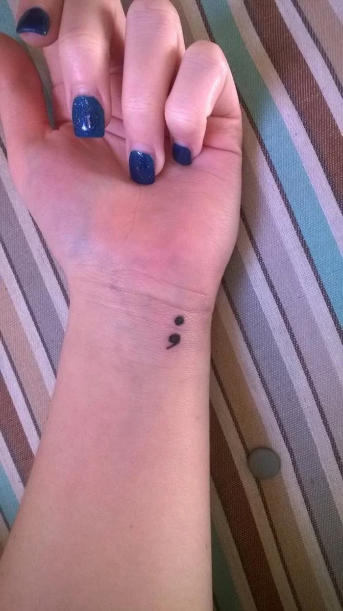 suicide awareness tattoo, of a small black semicolon, on a person's wrist, hand with long nails, painted in dark blue sparkly nail polish