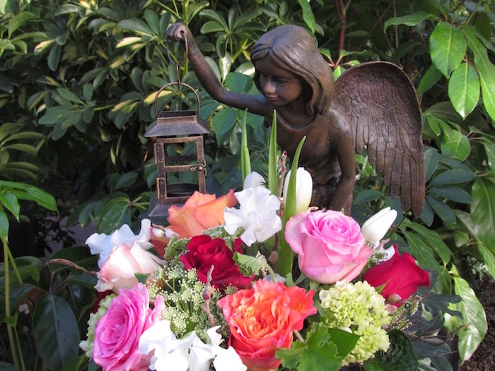 bouquet with pink, red and orange roses, and several white flowers, placed in a garden, near a small angel statue, holding a lantern, fairy garden images, foliage in the background