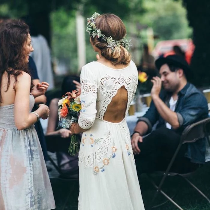 embroidered white gown, with lace inserts, cutout back and floral motives, casual beach wedding dresses, worn by bride with light brunette hair, in a messy bun, delicate flower crown