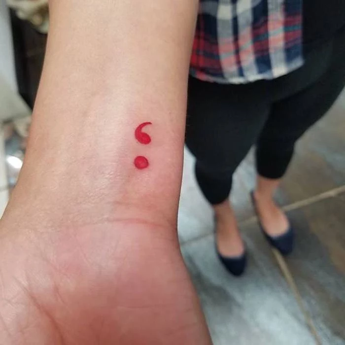 red semicolon tattoo, delicate minimalistic design, on a slim wrist, semicolon meaning and ideas, woman dressed in black leggings, ballerina flats and a plaid shirt