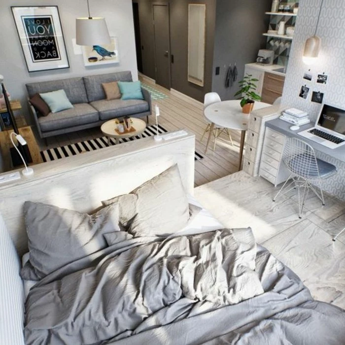 messy bed in white and gray, near blue-gray desk and matching chair, light wooden floor, grey sofa and white round table, small apartment ideas, framed artworks and shelves