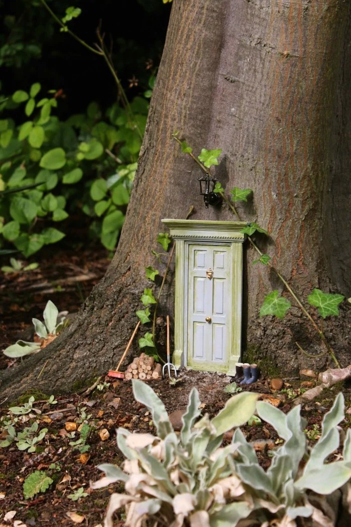 door in vintage style, made in miniature size, with golden knocker and door knob, attached to the trunk of a tree, near two pairs of tiny rain boots, and gardening gear