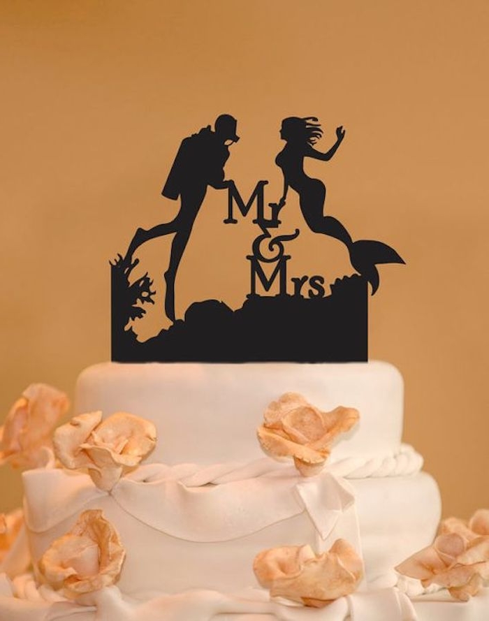 black cake topper, depicting the silhouettes of a mermaid and a diver, along with the words M & Mrs, placed on a white cake with peach-colored fondant flowers