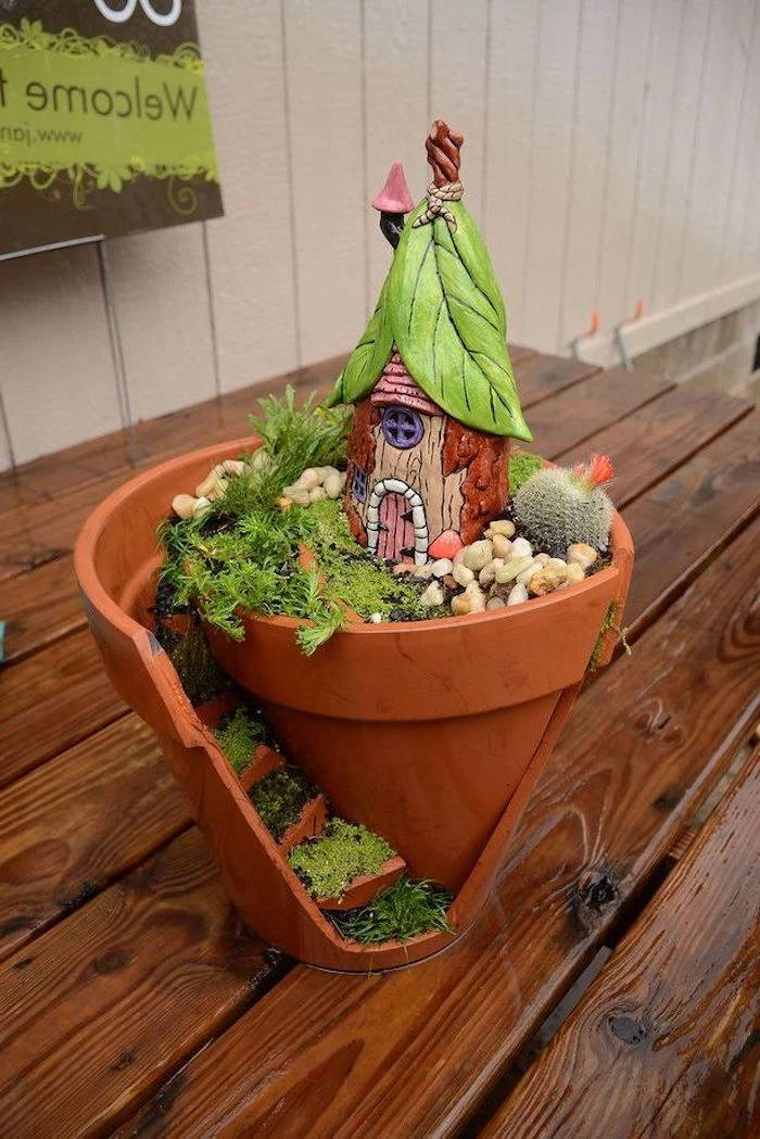 broken pot transformed into a diy fairy house, decorated with faux stairs, green moss and pebbles, cactus and a hand-painted ceramic house figurine