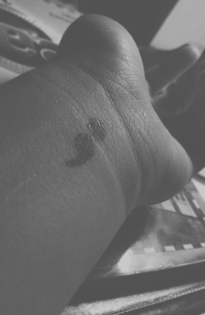 grayscale faded photo, of a person's wrist, showing a black semicolon tattoo, or suicide awareness tattoo, outstretched hand, resting on a table