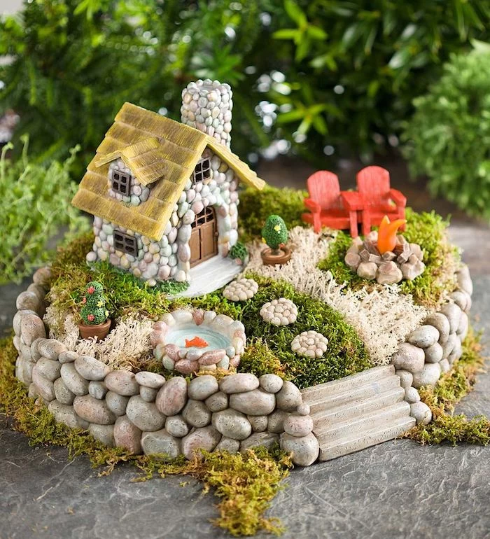 couple of red miniature armchairs, on green and pale beige moss, inside a miniature fairy garden, with tiny house and goldfish pond