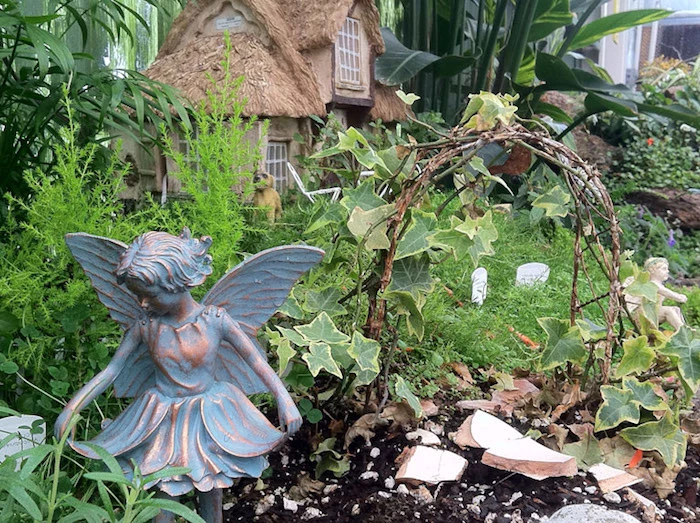 antique metal statue of a fairy, placed near a small arch, made from twigs and ivy, garden with various plants, and a miniature house in the background, fairy garden pictures