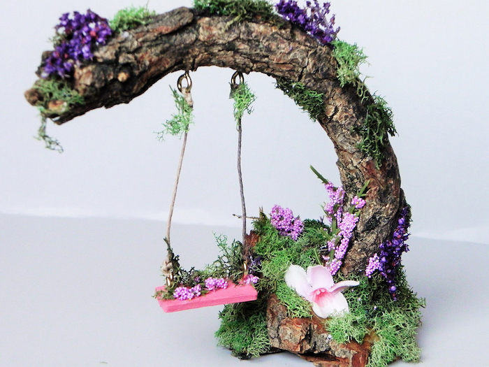 pink miniature swing, hanging from a dried branch, fairy garden pictures, decorated with purple and pink flowers, and green moss