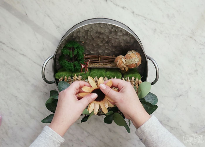fake sunflower being added to a spring wreath, made from aluminium cooking pot, containing fairy garden, little figurines and wheat stalks, moss and dried mushrooms