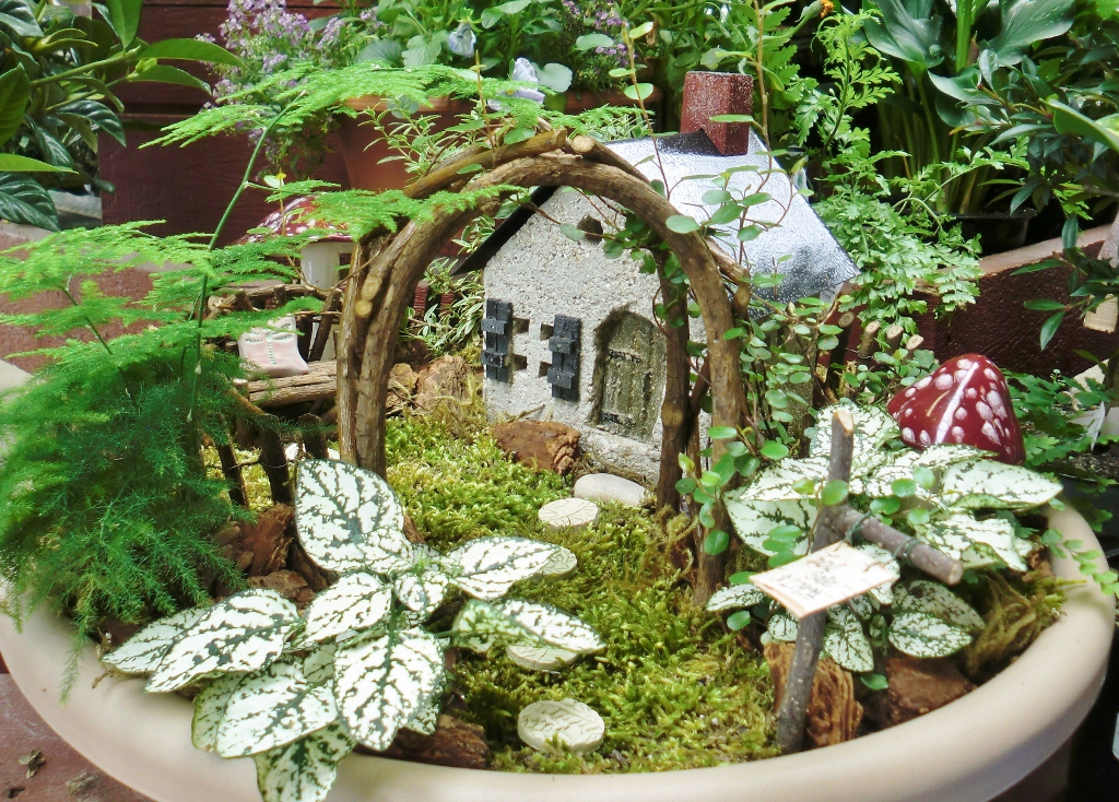 arch made of tiny woven branches, near ferns and other green plants, in a moss covered pot, decorated with a tiny fairy house, and mushroom figurines