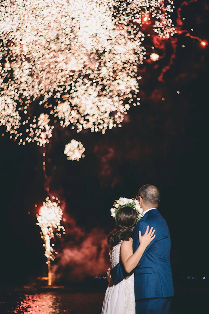 hugging bride in embroidered gown and groom in blue suit, watching fireworks over the sea, in the night sky, beach wedding venues