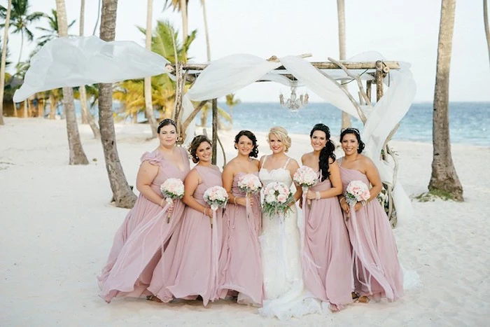 rosy ash pink, floaty bridesmaid dresses, on five women, each holding a small bouquet, standing next to a smiling bride, sea in the background