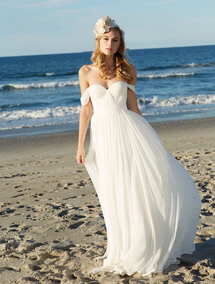 floaty white gown, with slouching shoulder straps, beach wedding dresses, worn by blonde woman with curled hair, and floral hair ornament in cream, standing on a sandy beach