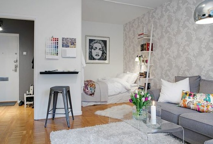 artwork of madonna in black and white, hanging over a small bed, in one-room flat, with gray sofa and pale gray rug, how to decorate a studio apartment, laminate floor and glass coffee table