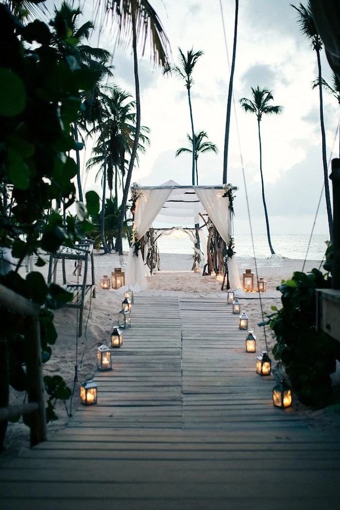 tall palm trees, on a sandy white shore, wooden boards leading to a simple wedding tent, made from wooden poles and white cloth, beach wedding venues, lots of lit lanterns