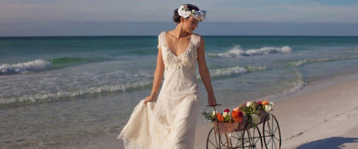 cart with red, white and yellow flowers, pulled by brunette woman, wearing white vintage style, embroidered bridal gown, and a flower crown, beach weddings in florida, sea waves in the background