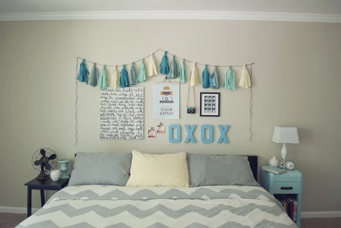 tassels in cream and three shades of blue, attached to a string, hanging over posters and decorations, on wall above a double bed in gray and cream