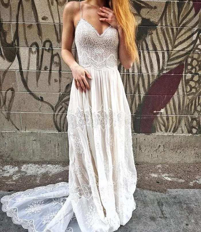 copper blonde woman, dressed in an embroidered, maxi wedding dress, in ivory and white, with long lace hem, standing in front of a graffitied wall