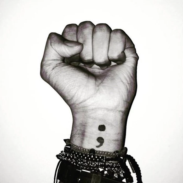 photo of a hand shaped into a fist, in black and white, several bracelets with studs and chains, wrist suicide awareness tattoo of a semicolon