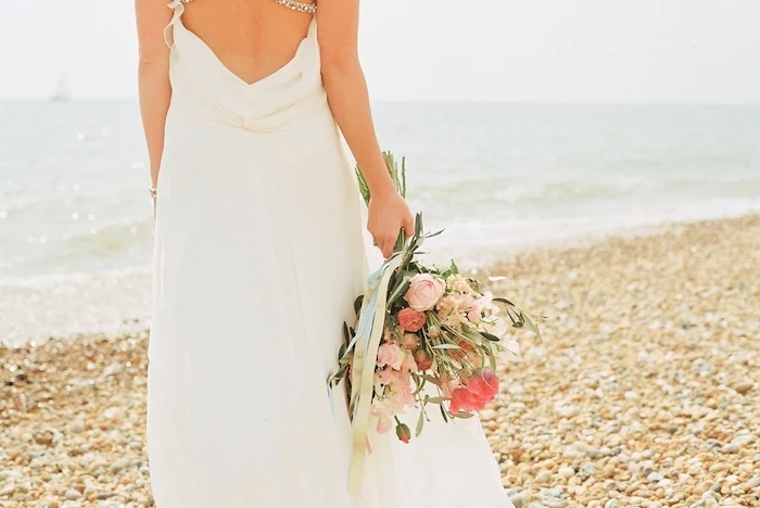 wedding dresses for beach wedding, white grecian style gown, with slouched back, worn by slim woman, standing on a pebbled beach, holding bouquet of pale and saturated pink flowers