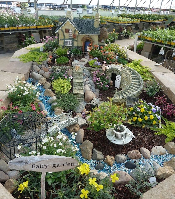 winding stone stairs, of a tiny size, near black wire gazebo, little white bridge, a decorative house, and other ornaments, diy fairy house, in a garden with different flowers, placed inside a large greenhouse