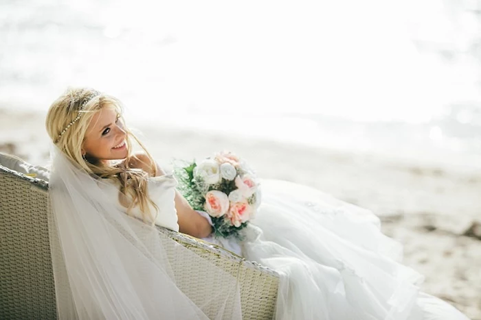 long sheer white veil, worn by smiling blonde bride, sitting on an ivory-colored wicker settee, placed on a sandy beach, wedding dresses for beach wedding, bouquet of white and pale peach flowers