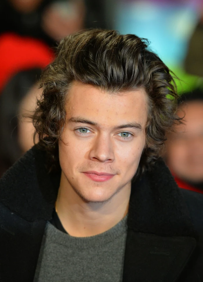 former one direction member, harry styles with wavy, messy brunette hair, bangs slicked back, curly hairstyles, wearing gray jumper, and black woollen coat with large collar