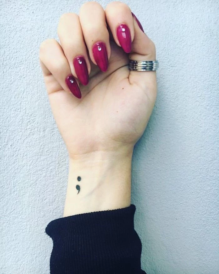 pinkish-purple nail polish, on long oval nails, pale white hand, with silver thumb ring, small black semicolon tattoo on the wrist, black jumper sleeve