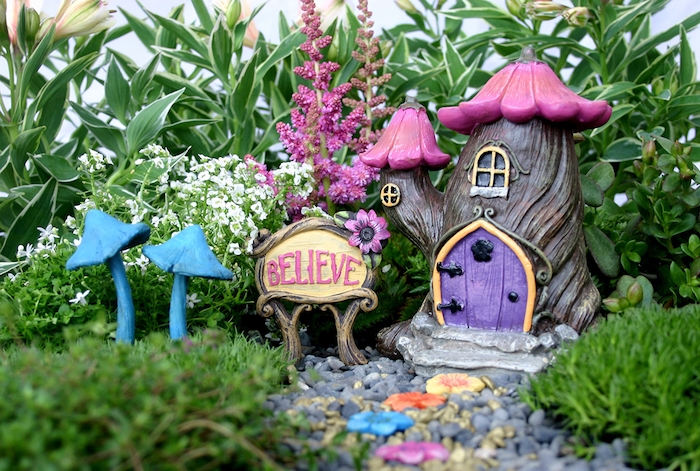 believe written with pink letters, on a board next to a fairy house ornament, made to look like a small tree, with pink flowers roof, two blue mushroom figurines, and many green plants