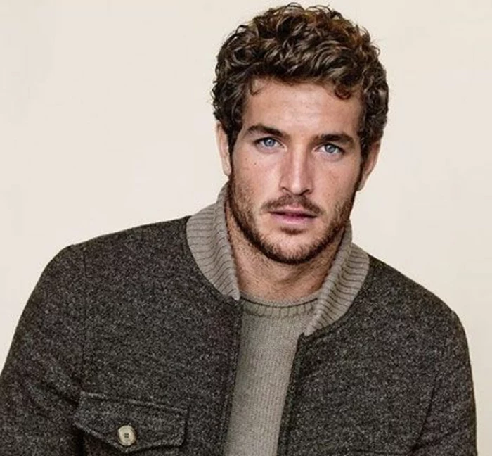 dynamic curly hairstyle, on a brunette man with blue eyes, wearing dark gray jacket, over khaki grey knitted jumper 