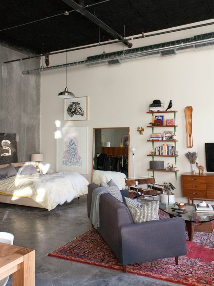 black ceiling with metal pipes and details, in open-plan industrial style home, how to decorate a studio apartment, double bed and dark gray sofa, red patterned kilim rug, bookshelves and decorations