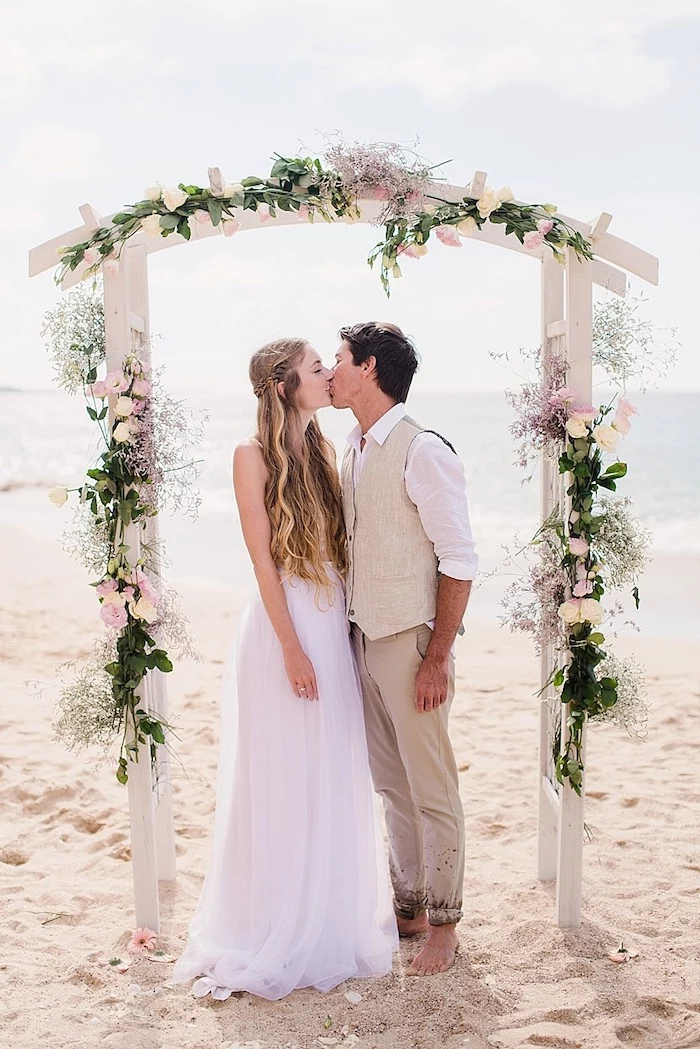 young woman with wavy highlighted dark blonde hair, wearing a white maxi dress with tulle skirt, kissing a man in pale beige suit, under a wedding arch, beach wedding venues near the sea