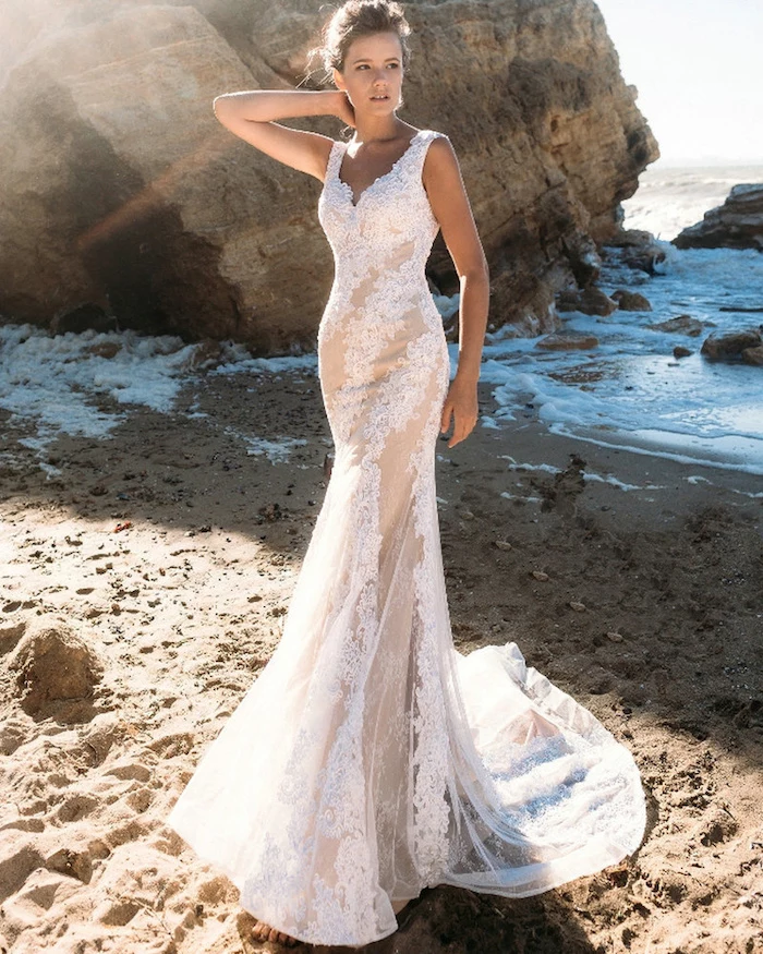 classical mermaid lace beach wedding dress, in white and cream, worn by tall and slim young woman, with brunette hair in a messy hairdo, boulders sand and sea