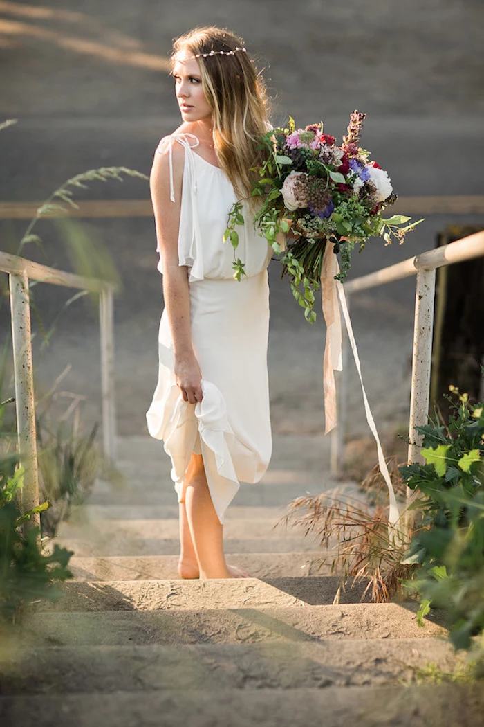 simple white grecian wedding gown, beach wedding dresses, worn by young woman, with delicate flower crown, holding a large bouquet, standing on sandy steps