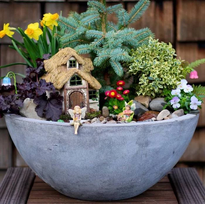 fir branch and many different flowers, and green plants, inside a large gray bowl, with pebbles and fairy figurines, pansies and daffodils